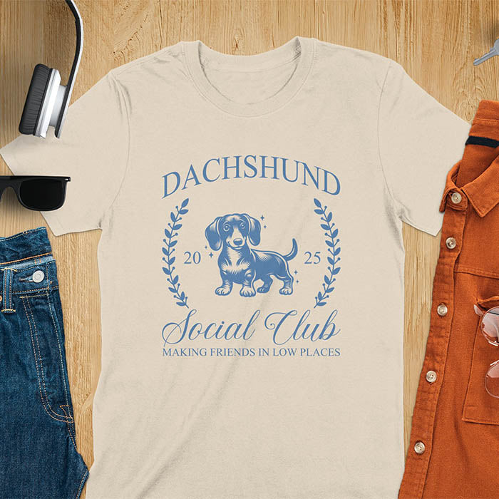 Sand color T-shirt with a playful dachshund graphic, laurel branches, and text reading "Dachshund Social Club," "EST 2024," and "Making Friends in Low Places." Vintage-inspired design perfect for dog lovers.