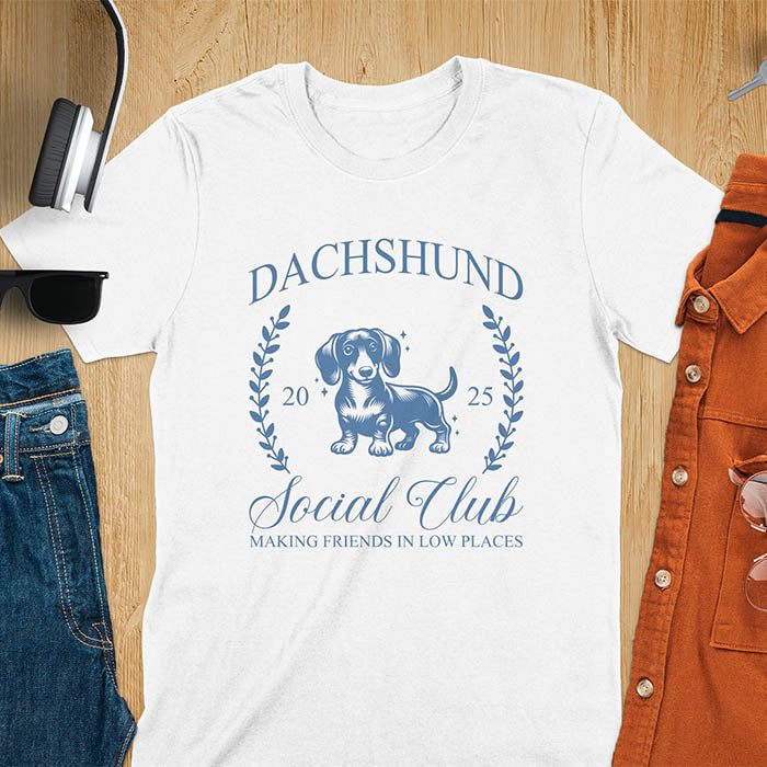 White T-shirt with a playful dachshund graphic, laurel branches, and text reading "Dachshund Social Club," "EST 2024," and "Making Friends in Low Places." Vintage-inspired design perfect for dog lovers.