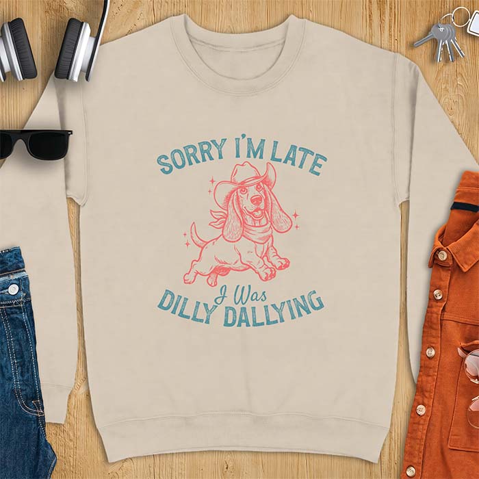 Sand sweatshirt with a graphic of a basset hound wearing a cowboy hat and bandana trotting with sparkles around it and the text Sorry I'm Late I Was Dilly Dallying