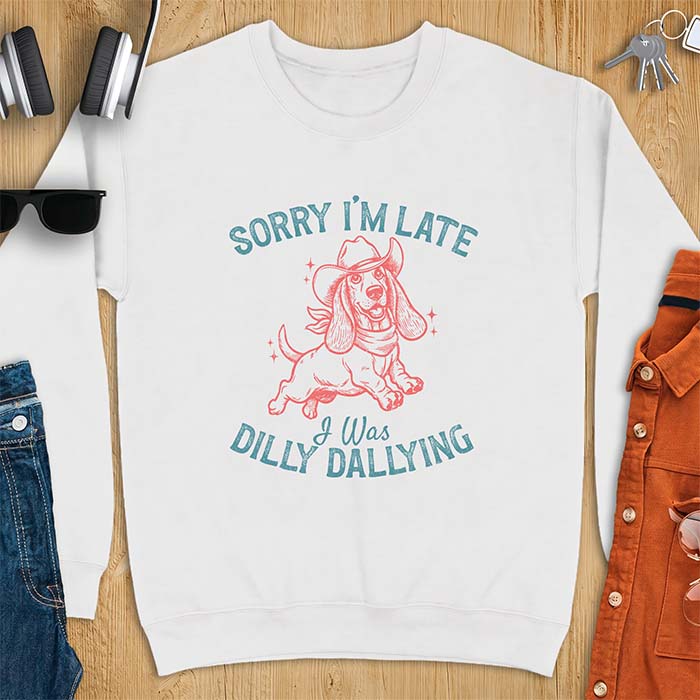 White sweatshirt with a graphic of a basset hound wearing a cowboy hat and bandana trotting with sparkles around it and the text Sorry I'm Late I Was Dilly Dallying