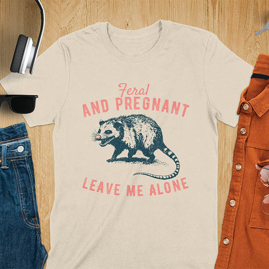 A sand color t-shirt featuring An upset-looking opossum showing its fangs. Text reads Feral and Pregnant and Leave Me Alone.