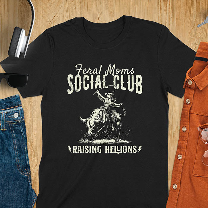 A black color t-shirt with a cowgirl riding a bucking bull. Text: Feral Moms SOCIAL CLUB above and Raising HELLIONS below. This unhinged t-shirts design features a cowgirl mom on a rodeo bucking bull, a weird meme shirt for a feral mom gift. A retro inspired design, one of those shirts that go hard, a mama outfit or feral wife gift. A mom gift or gift for new mom for a western mama.