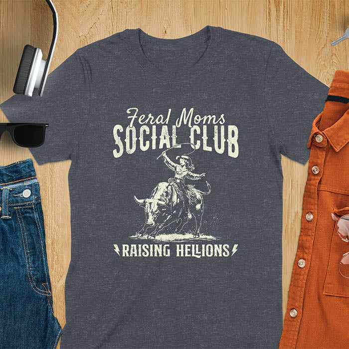 A dark heather t-shirt with a cowgirl riding a bucking bull. Text: Feral Moms SOCIAL CLUB above and Raising HELLIONS below. This unhinged t-shirts design features a cowgirl mom on a rodeo bucking bull, a weird meme shirt for a feral mom gift. A retro inspired design, one of those shirts that go hard, a mama outfit or feral wife gift. A mom gift or gift for new mom for a western mama.
