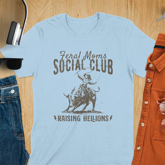 A light blue t-shirt with a cowgirl riding a bucking bull. Text: Feral Moms SOCIAL CLUB above and Raising HELLIONS below. This unhinged t-shirts design features a cowgirl mom on a rodeo bucking bull, a weird meme shirt for a feral mom gift. A retro inspired design, one of those shirts that go hard, a mama outfit or feral wife gift. A mom gift or gift for new mom for a western mama.