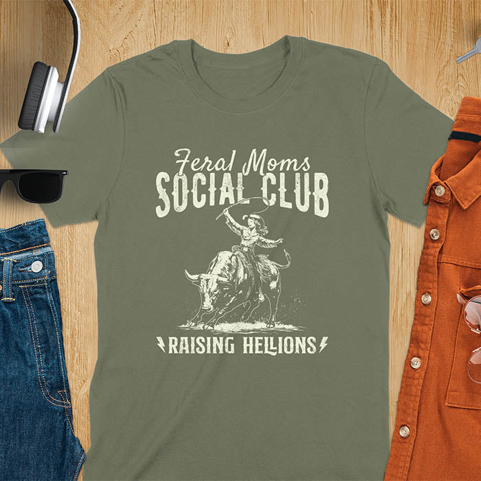A military green t-shirt with a cowgirl riding a bucking bull. Text: Feral Moms SOCIAL CLUB above and Raising HELLIONS below. This unhinged t-shirts design features a cowgirl mom on a rodeo bucking bull, a weird meme shirt for a feral mom gift. A retro inspired design, one of those shirts that go hard, a mama outfit or feral wife gift. A mom gift or gift for new mom for a western mama.