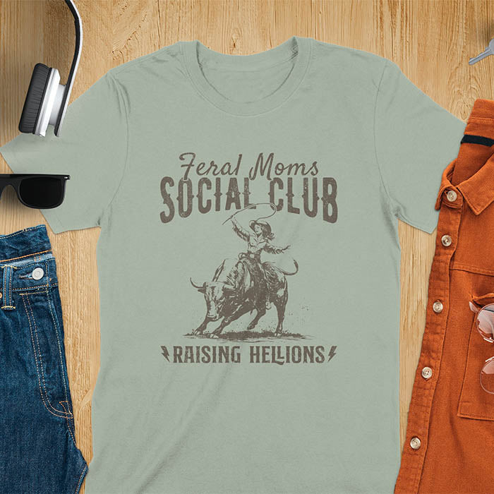 A sage color t-shirt with a cowgirl riding a bucking bull. Text: Feral Moms SOCIAL CLUB above and Raising HELLIONS below. This unhinged t-shirts design features a cowgirl mom on a rodeo bucking bull, a weird meme shirt for a feral mom gift. A retro inspired design, one of those shirts that go hard, a mama outfit or feral wife gift. A mom gift or gift for new mom for a western mama.