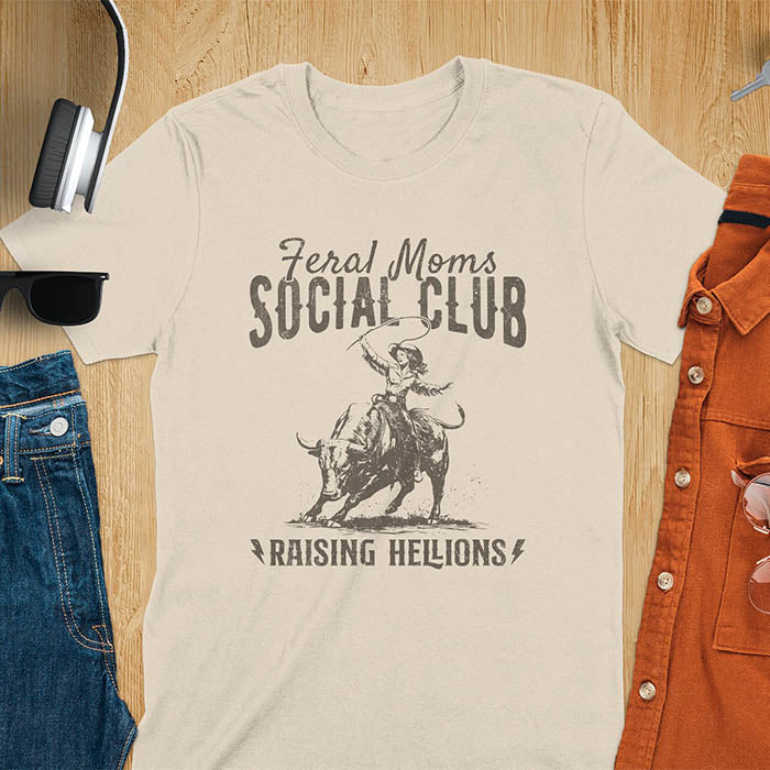A sand color t-shirt with a cowgirl riding a bucking bull. Text: Feral Moms SOCIAL CLUB above and Raising HELLIONS below. This unhinged t-shirts design features a cowgirl mom on a rodeo bucking bull, a weird meme shirt for a feral mom gift. A retro inspired design, one of those shirts that go hard, a mama outfit or feral wife gift. A mom gift or gift for new mom for a western mama.