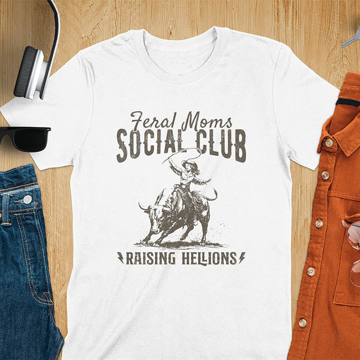 A white color t-shirt with a cowgirl riding a bucking bull. Text: Feral Moms SOCIAL CLUB above and Raising HELLIONS below. This unhinged t-shirts design features a cowgirl mom on a rodeo bucking bull, a weird meme shirt for a feral mom gift. A retro inspired design, one of those shirts that go hard, a mama outfit or feral wife gift. A mom gift or gift for new mom for a western mama.