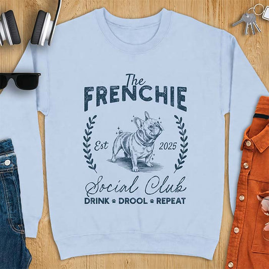 Light Blue sweatshirt with a vintage badge graphic of a French Bulldog wearing a collar and tag framed by a laurel wreath, and the text The Frenchie, Est 2025, Social Club, Drink Drool Repeat