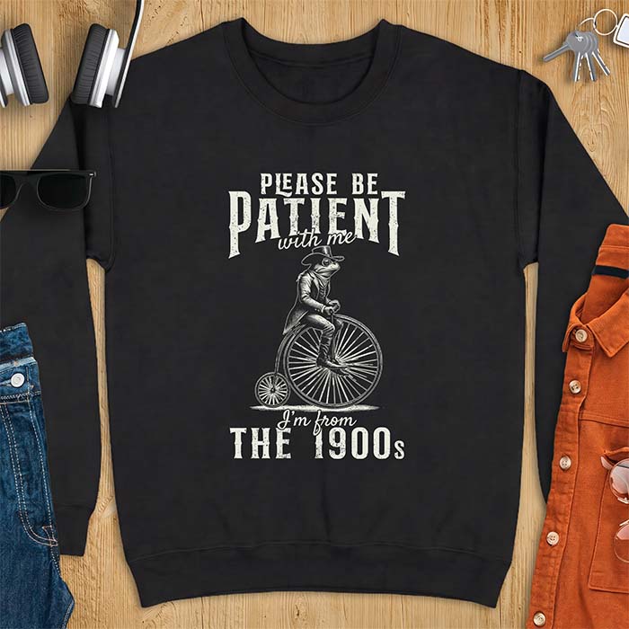 Black sweatshirt with a graphic of a frog dressed in a Victorian coat, vest, top hat and monocle riding a penny farthing bicycle and the text Please Be Patient With Me I'm From The 1900s