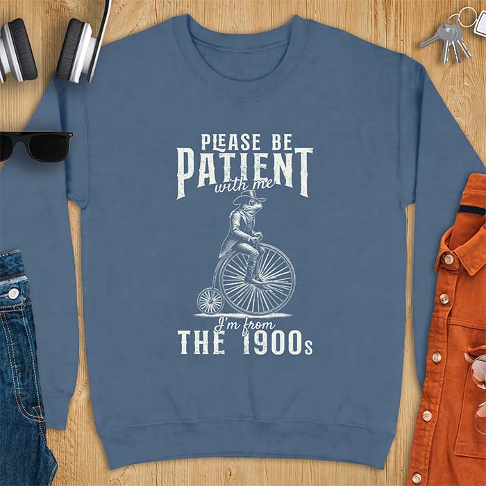 Indigo Blue sweatshirt with a graphic of a frog dressed in a Victorian coat, vest, top hat and monocle riding a penny farthing bicycle and the text Please Be Patient With Me I'm From The 1900s