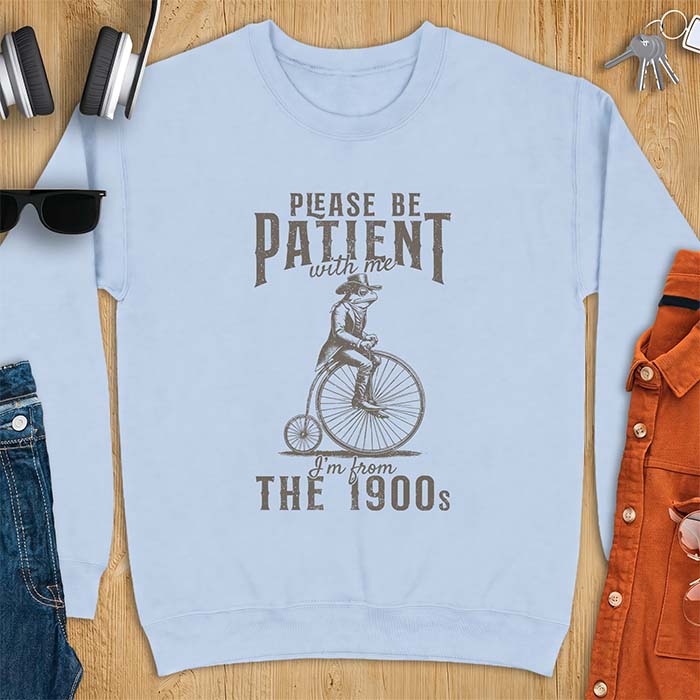 Light Blue sweatshirt with a graphic of a frog dressed in a Victorian coat, vest, top hat and monocle riding a penny farthing bicycle and the text Please Be Patient With Me I'm From The 1900s