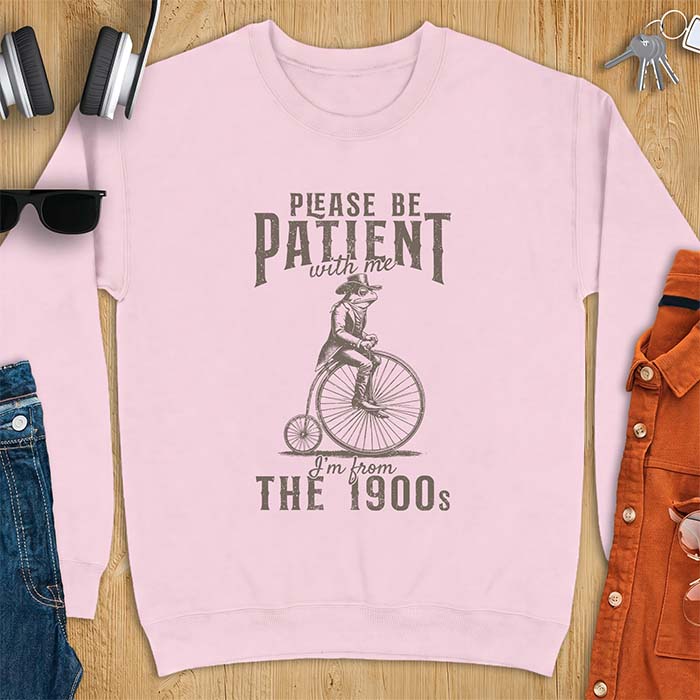Light Pink sweatshirt with a graphic of a frog dressed in a Victorian coat, vest, top hat and monocle riding a penny farthing bicycle and the text Please Be Patient With Me I'm From The 1900s