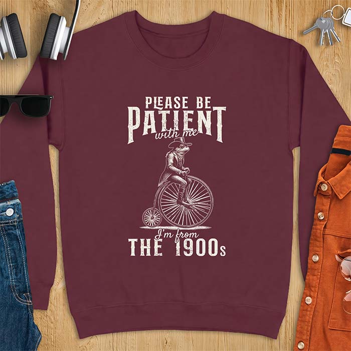 Maroon sweatshirt with a graphic of a frog dressed in a Victorian coat, vest, top hat and monocle riding a penny farthing bicycle and the text Please Be Patient With Me I'm From The 1900s