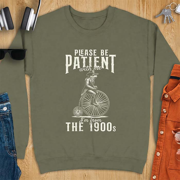 Military Green sweatshirt with a graphic of a frog dressed in a Victorian coat, vest, top hat and monocle riding a penny farthing bicycle and the text Please Be Patient With Me I'm From The 1900s