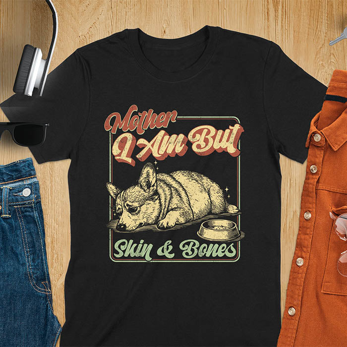 Chubby Corgi lying next to an empty bowl with a sulky expression, with the text Mother I Am But Skin and Bones in retro typography on a black t-shirt