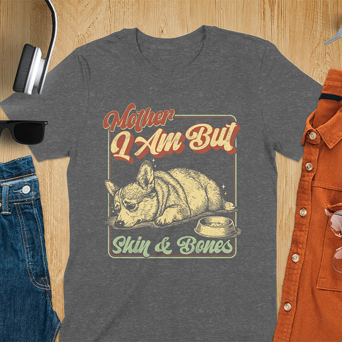 Chubby Corgi lying next to an empty bowl with a sulky expression, with the text Mother I Am But Skin and Bones in retro typography on a dark heather t-shirt