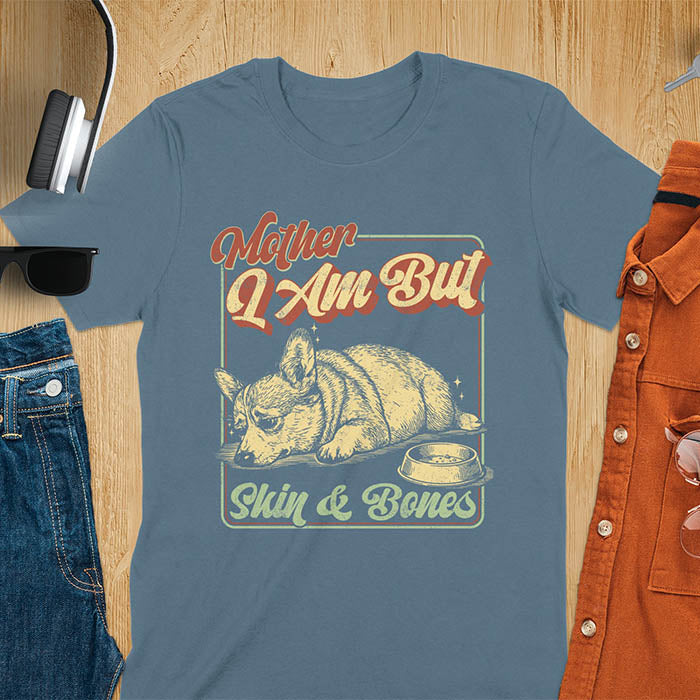Chubby Corgi lying next to an empty bowl with a sulky expression, with the text Mother I Am But Skin and Bones in retro typography on a indigo blue t-shirt