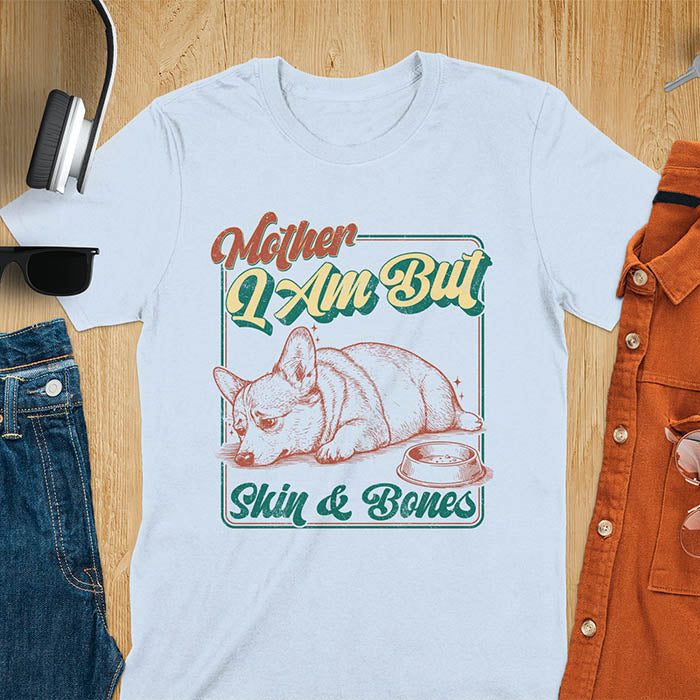 Chubby Corgi lying next to an empty bowl with a sulky expression, with the text Mother I Am But Skin and Bones in retro typography on a light blue t-shirt
