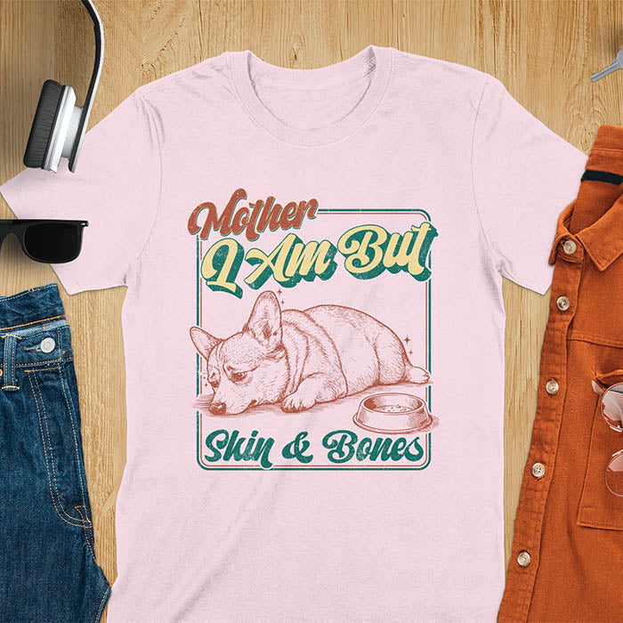 Chubby Corgi lying next to an empty bowl with a sulky expression, with the text Mother I Am But Skin and Bones in retro typography on a light pink t-shirt