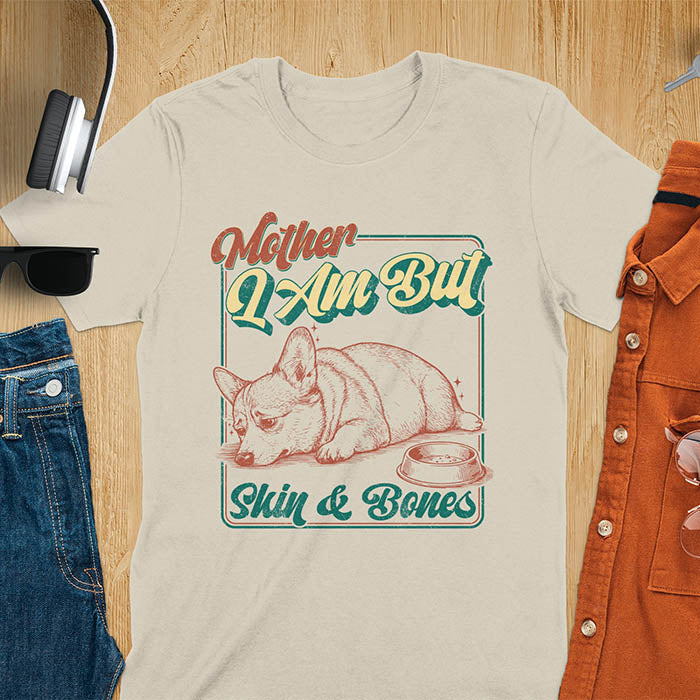 Chubby Corgi lying next to an empty bowl with a sulky expression, with the text Mother I Am But Skin and Bones in retro typography on a sand color t-shirt