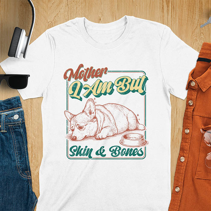Chubby Corgi lying next to an empty bowl with a sulky expression, with the text Mother I Am But Skin and Bones in retro typography on a white t-shirt