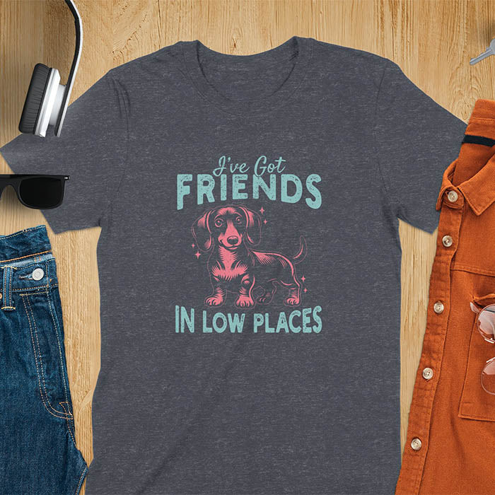 This T-shirt features a vintage-style line art of a happy dachshund looking straight at you with its tail held high. The top text reads, I've got friends, and below it says, in low places. Overall in a retro color palette with a distressed texture, on a dark heather tee.