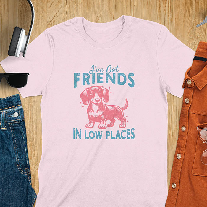 This T-shirt features a vintage-style line art of a happy dachshund looking straight at you with its tail held high. The top text reads, I've got friends, and below it says, in low places. Overall in a retro color palette with a distressed texture, on a light pink tee.