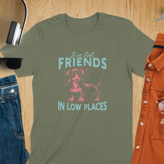This T-shirt features a vintage-style line art of a happy dachshund looking straight at you with its tail held high. The top text reads, I've got friends, and below it says, in low places. Overall in a retro color palette with a distressed texture, on a military green tee.