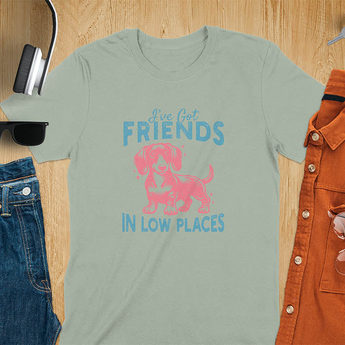This T-shirt features a vintage-style line art of a happy dachshund looking straight at you with its tail held high. The top text reads, I've got friends, and below it says, in low places. Overall in a retro color palette with a distressed texture, on a sage green tee.