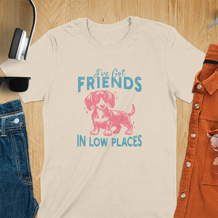 This T-shirt features a vintage-style line art of a happy dachshund looking straight at you with its tail held high. The top text reads, I've got friends, and below it says, in low places. Overall in a retro color palette with a distressed texture, on a sand color tee.