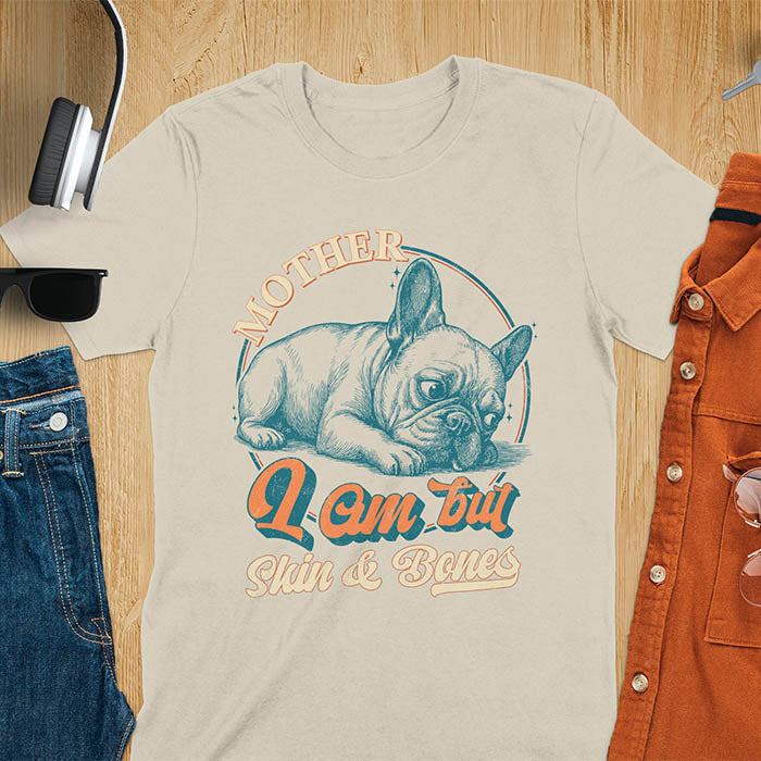 French Bulldog lying down with a sulky expression, with the text Mother I Am But Skin and Bones in retro typography on a sand color t-shirt