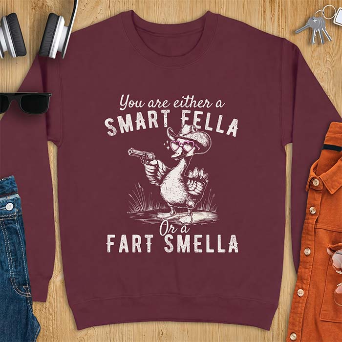 Maroon sweatshirt with a cowboy goose illustration wearing a hat and pink heart shaped sunglasses, holding a revolver, and the text You Are Either A Smart Fella Or A Fart Smella