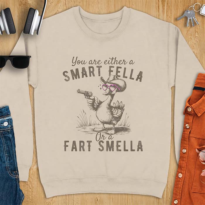 Sand sweatshirt with a cowboy goose illustration wearing a hat and pink heart shaped sunglasses, holding a revolver, and the text You Are Either A Smart Fella Or A Fart Smella