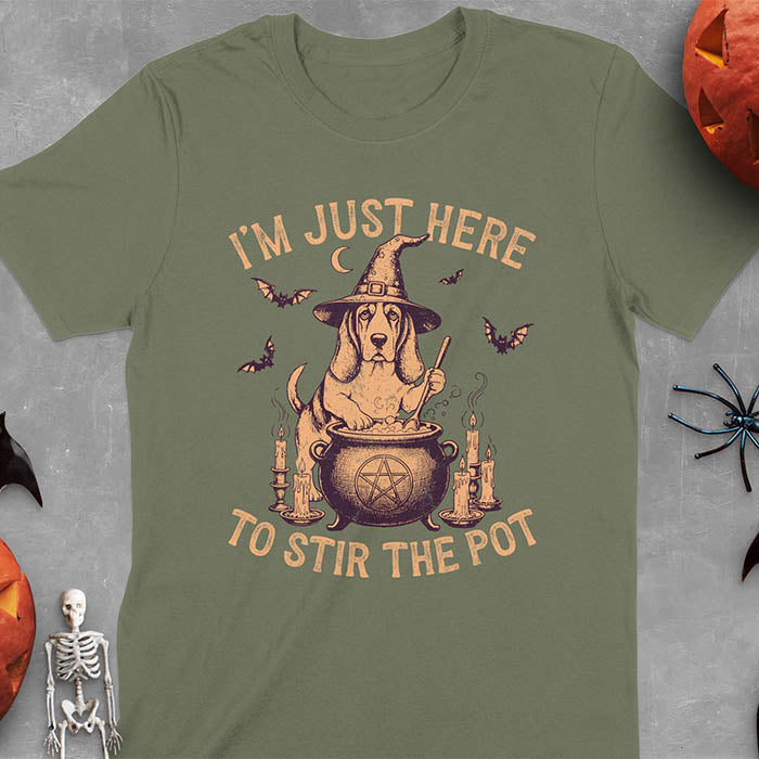Military Green t-shirt featuring Halloween Basset Hound witch stirring cauldron with pentagram, lit candles on sides, crescent moon and bats in background, wearing pointed witch hat with text I'm Just Here To Stir The Pot