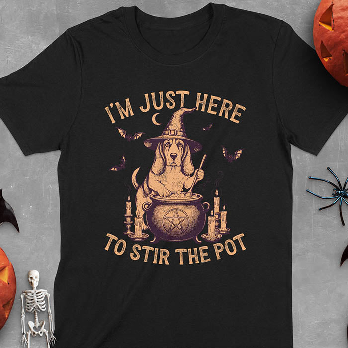 Black t-shirt featuring Halloween Basset Hound witch stirring cauldron with pentagram, lit candles on sides, crescent moon and bats in background, wearing pointed witch hat with text I'm Just Here To Stir The Pot