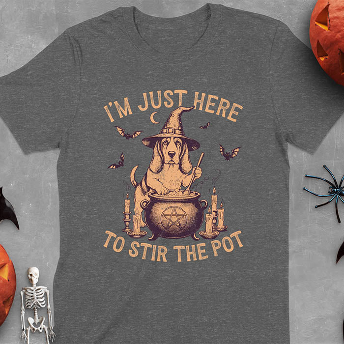 Dark heather t-shirt featuring Halloween Basset Hound witch stirring cauldron with pentagram, lit candles on sides, crescent moon and bats in background, wearing pointed witch hat with text I'm Just Here To Stir The Pot