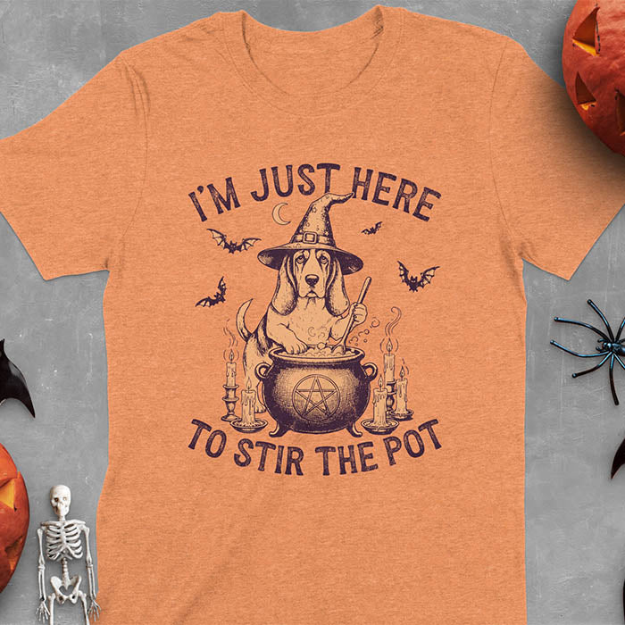 Heather Orange t-shirt featuring Halloween Basset Hound witch stirring cauldron with pentagram, lit candles on sides, crescent moon and bats in background, wearing pointed witch hat with text I'm Just Here To Stir The Pot
