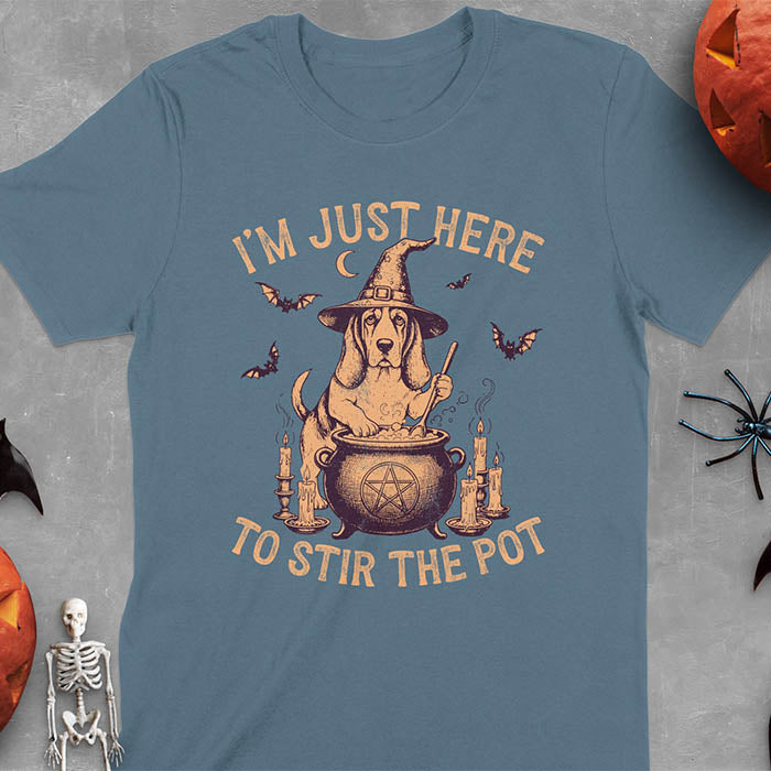 Indigo blue t-shirt featuring Halloween Basset Hound witch stirring cauldron with pentagram, lit candles on sides, crescent moon and bats in background, wearing pointed witch hat with text I'm Just Here To Stir The Pot