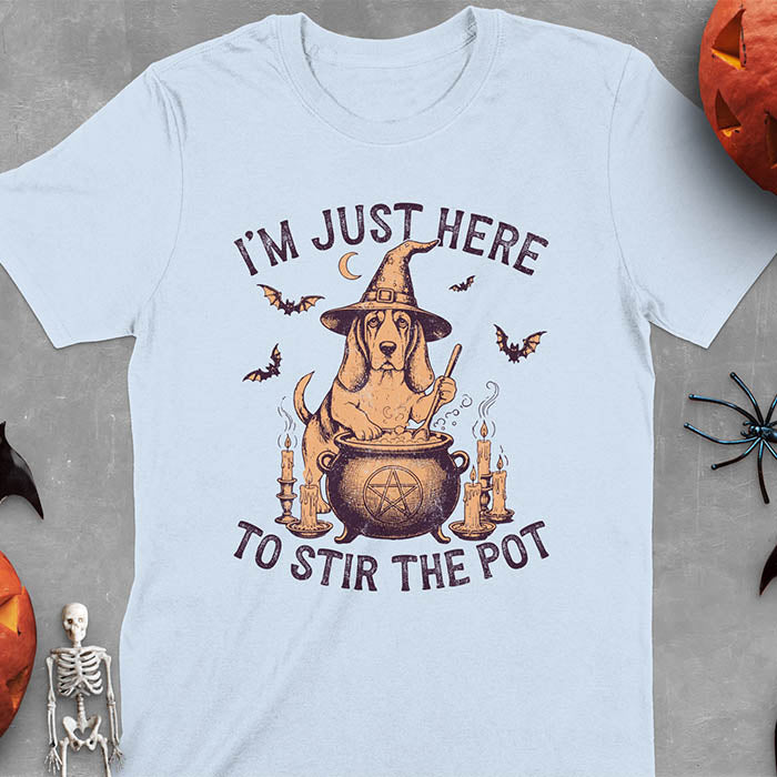Light blue t-shirt featuring Halloween Basset Hound witch stirring cauldron with pentagram, lit candles on sides, crescent moon and bats in background, wearing pointed witch hat with text I'm Just Here To Stir The Pot