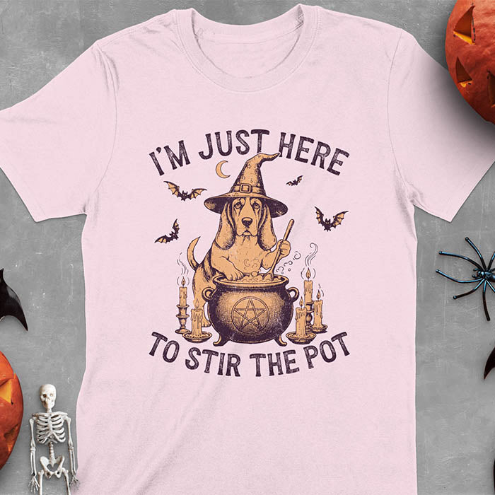 Light pink t-shirt featuring Halloween Basset Hound witch stirring cauldron with pentagram, lit candles on sides, crescent moon and bats in background, wearing pointed witch hat with text I'm Just Here To Stir The Pot