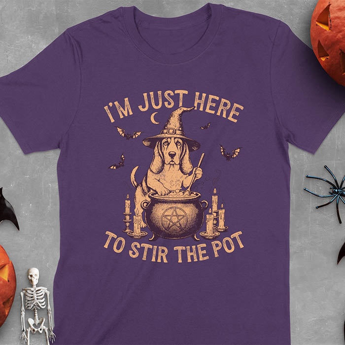 Purple t-shirt featuring Halloween Basset Hound witch stirring cauldron with pentagram, lit candles on sides, crescent moon and bats in background, wearing pointed witch hat with text I'm Just Here To Stir The Pot
