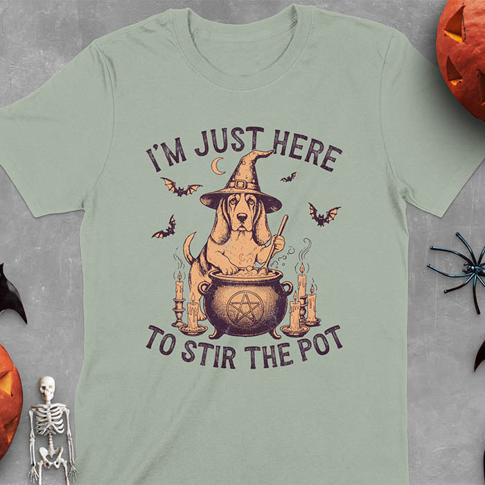 Sage green t-shirt featuring Halloween Basset Hound witch stirring cauldron with pentagram, lit candles on sides, crescent moon and bats in background, wearing pointed witch hat with text I'm Just Here To Stir The Pot