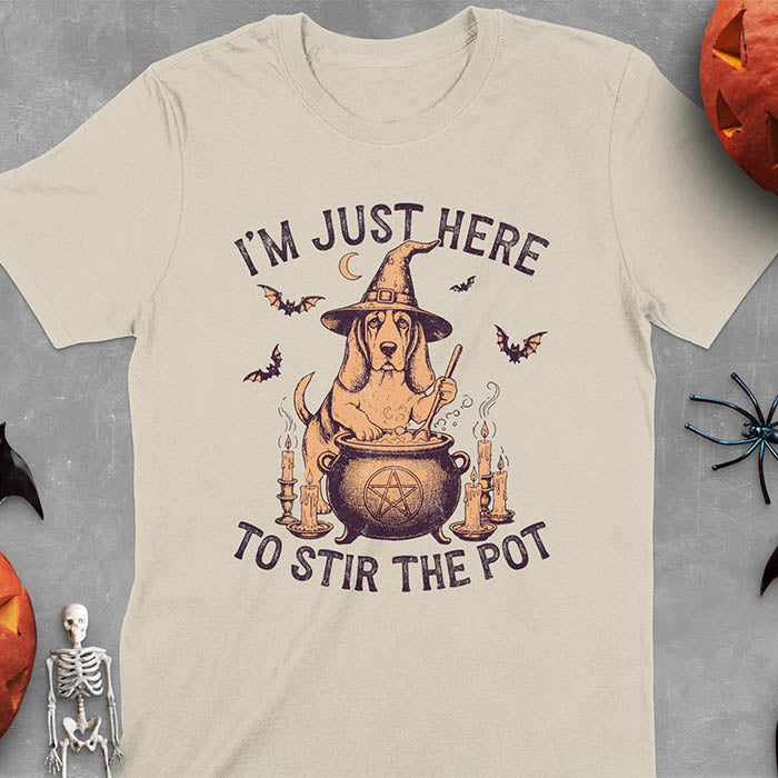 Sand color t-shirt featuring Halloween Basset Hound witch stirring cauldron with pentagram, lit candles on sides, crescent moon and bats in background, wearing pointed witch hat with text I'm Just Here To Stir The Pot