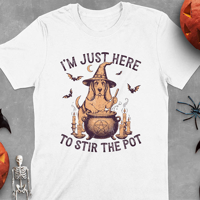 White t-shirt featuring Halloween Basset Hound witch stirring cauldron with pentagram, lit candles on sides, crescent moon and bats in background, wearing pointed witch hat with text I'm Just Here To Stir The Pot