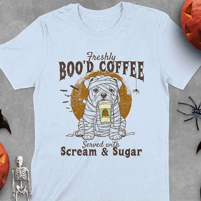 Light blue t-shirt featuring a mummy-wrapped bulldog holding a coffee cup with text 'Freshly BOO'D COFFEE Served with Scream & Sugar' with bats flying in background, full moon behind the bulldog, and a spider hanging from the coffee text