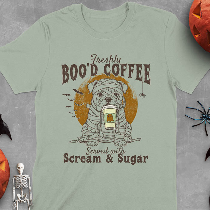 Sage green t-shirt featuring a mummy-wrapped bulldog holding a coffee cup with text 'Freshly BOO'D COFFEE Served with Scream & Sugar' with bats flying in background, full moon behind the bulldog, and a spider hanging from the coffee text