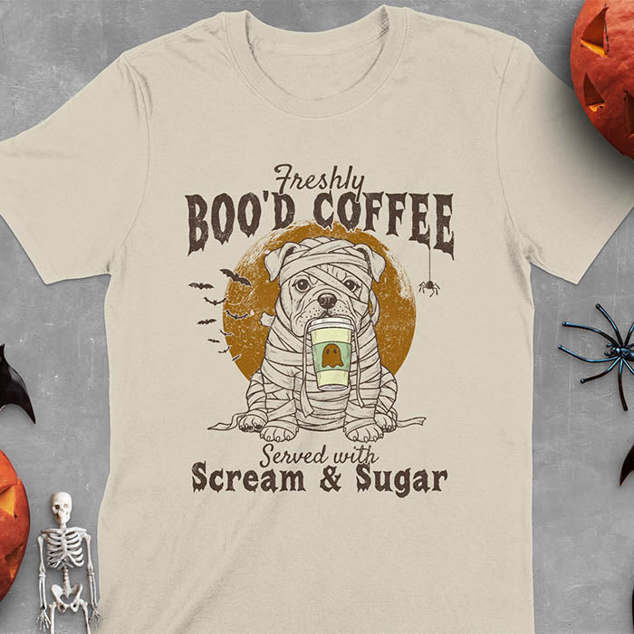 Sand colored t-shirt featuring a mummy-wrapped bulldog holding a coffee cup with text 'Freshly BOO'D COFFEE Served with Scream & Sugar' with bats flying in background, full moon behind the bulldog, and a spider hanging from the coffee text