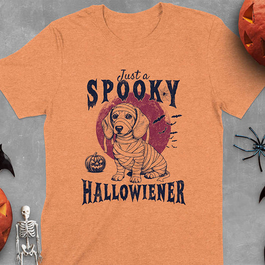 Heather Orange t-shirt featuring a cute dachshund puppy wrapped in mummy bandages with text 'Just a Spooky Hallowiener', jack-o'-lantern pumpkin, red moon and flying bats in background