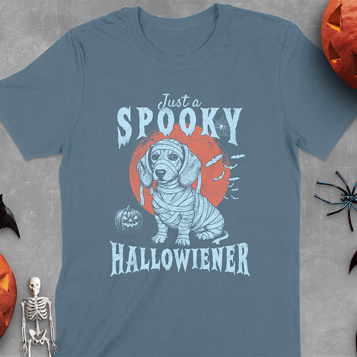 Indigo Blue t-shirt featuring a cute dachshund puppy wrapped in mummy bandages with text 'Just a Spooky Hallowiener', jack-o'-lantern pumpkin, red moon and flying bats in background